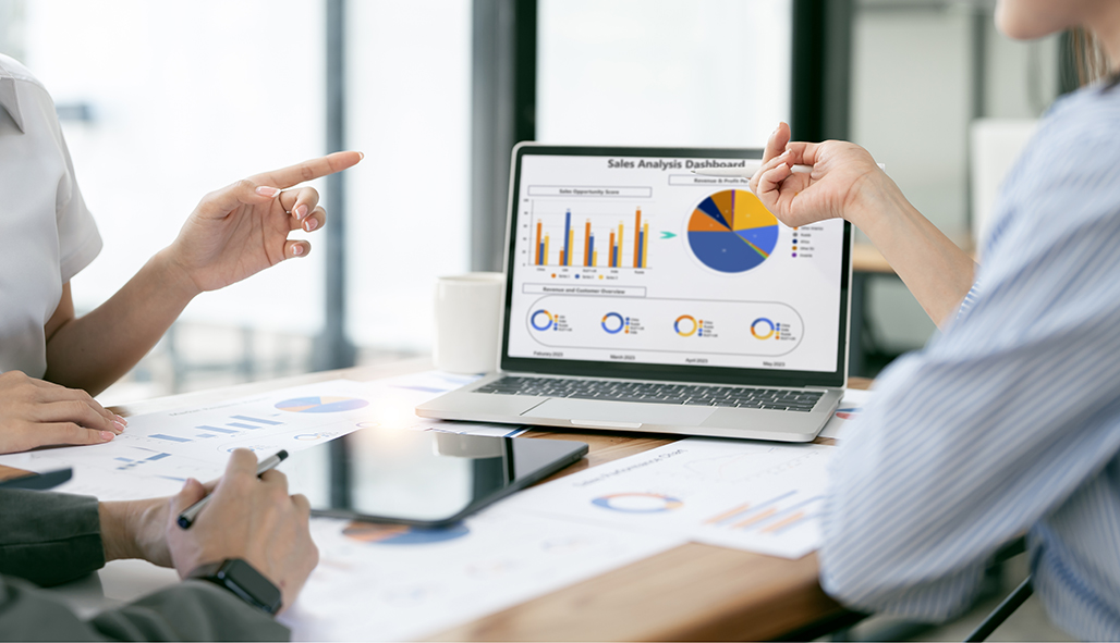 Two colleagues review a laptop screen showing a sales dashboard with bar charts and a pie chart on a bright desk.