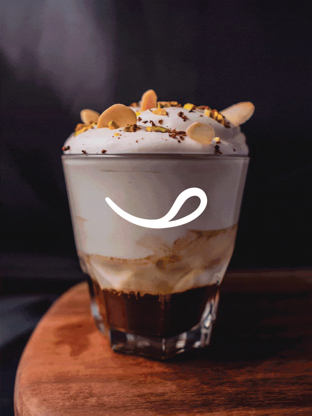 Glass tumbler with a layered coffee drink topped with whipped cream, chocolate crumbs, and yellow-green nut toppings, on a wooden surface.