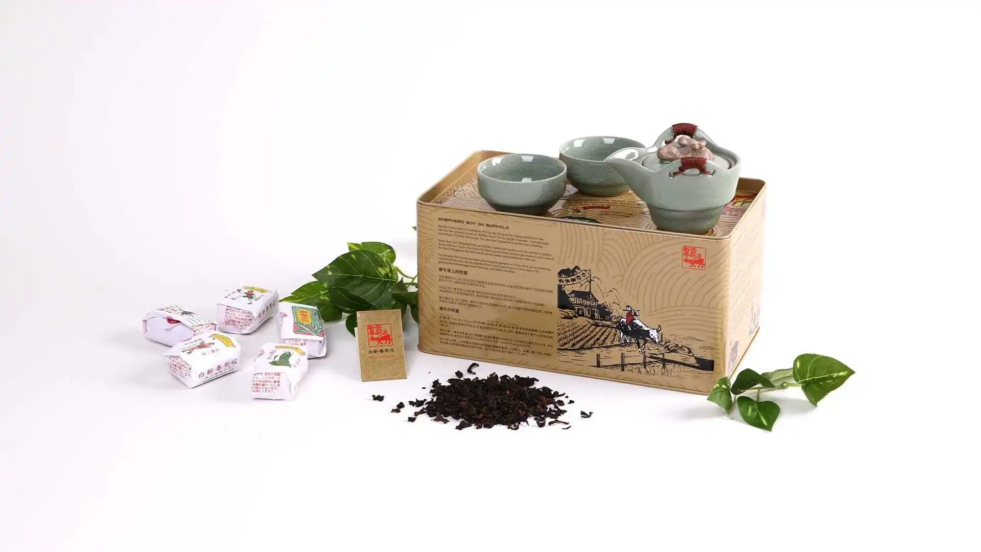 Pek Sin Choon tea set display featuring a illustrated tin box topped with a light green ceramic teapot and cups, surrounded by loose tea leaves, paper-wrapped tea packages, and green leaves.