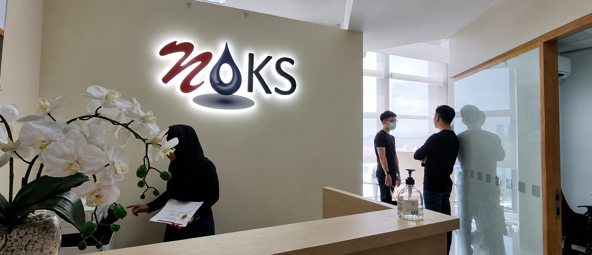 Reception area with a backlit 'NOKS' logo on a beige wall, white orchid plant on a wooden desk, and a woman in a hijab handing papers nearby.